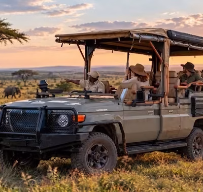 Safari jeep with four passengers in hats driving across a sunlit savanna, with distant grazing wildlife visible.