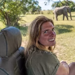 Smiling woman in a safari vehicle with elephants grazing in the background on a sunny day.